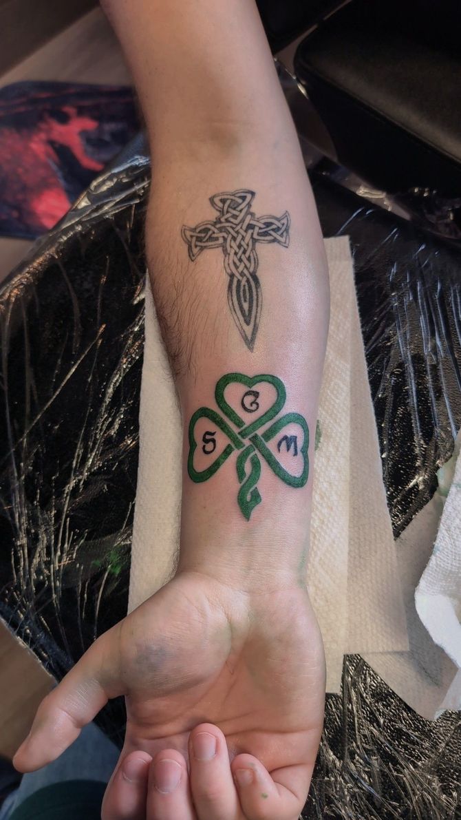 A person has a tattoo of a cross and a shamrock on their forearm.