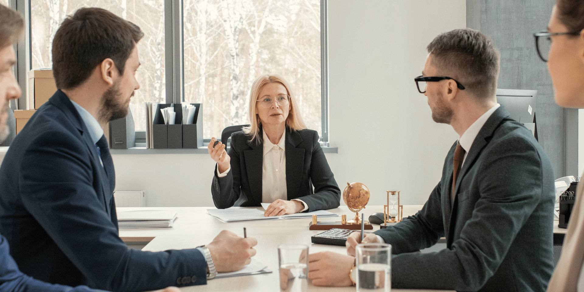 Business people in suits seated at a table, possibly in a meeting; one woman is speaking.