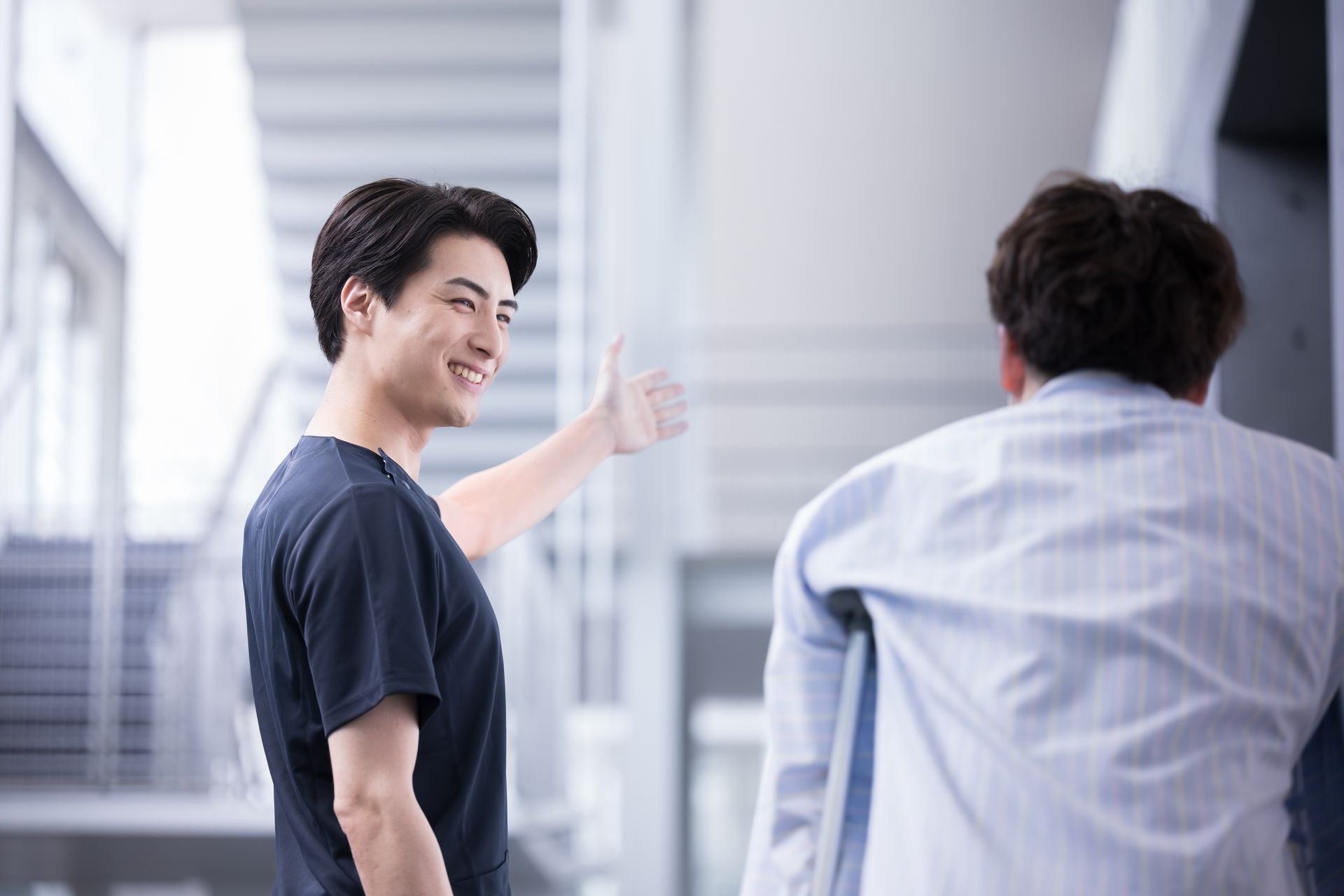 A medical professional in scrubs gestures toward a staircase, guiding a person toward an office or exam room.