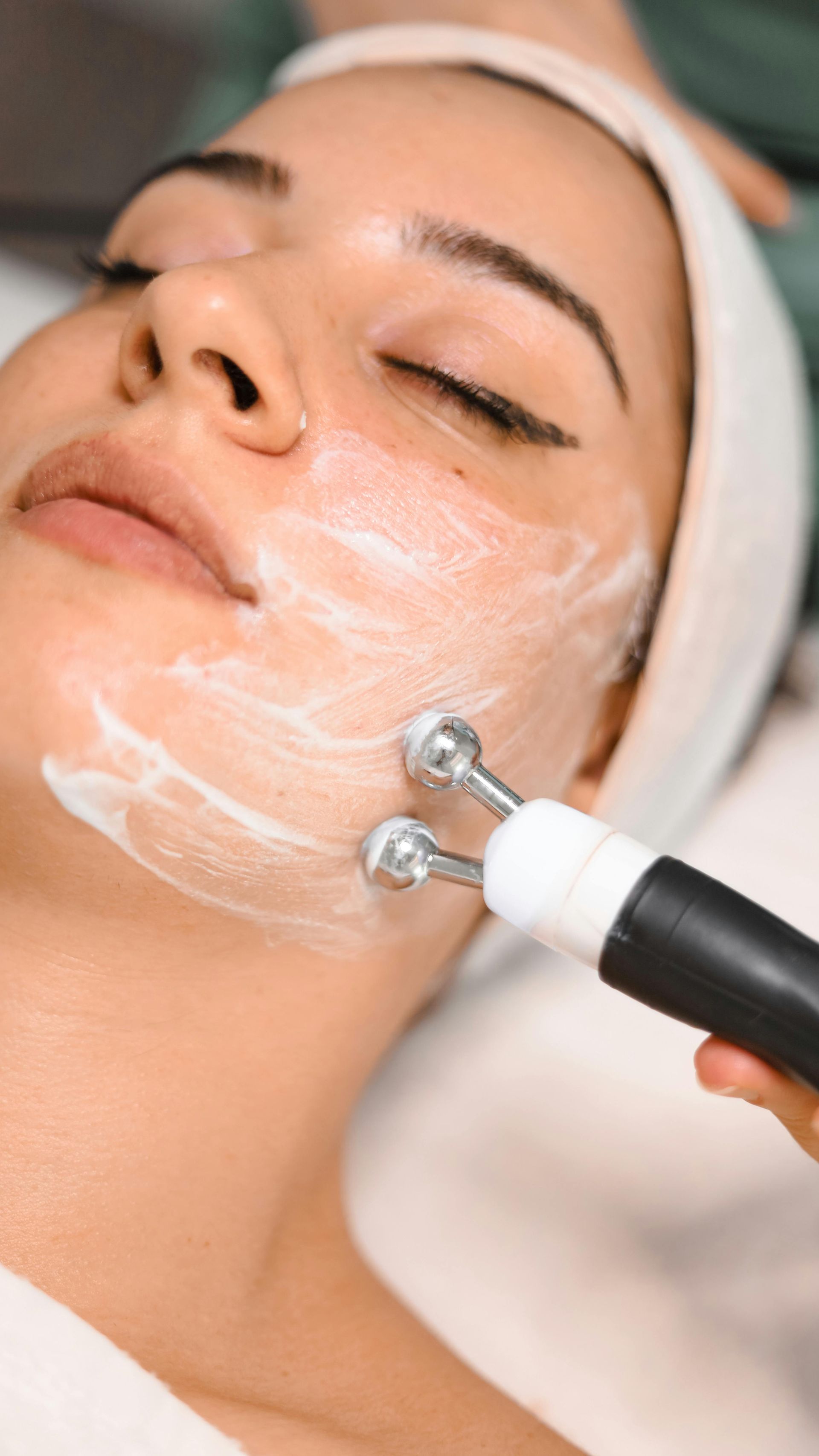 Woman receiving a facial treatment with a tool applying cream; lying down with closed eyes.
