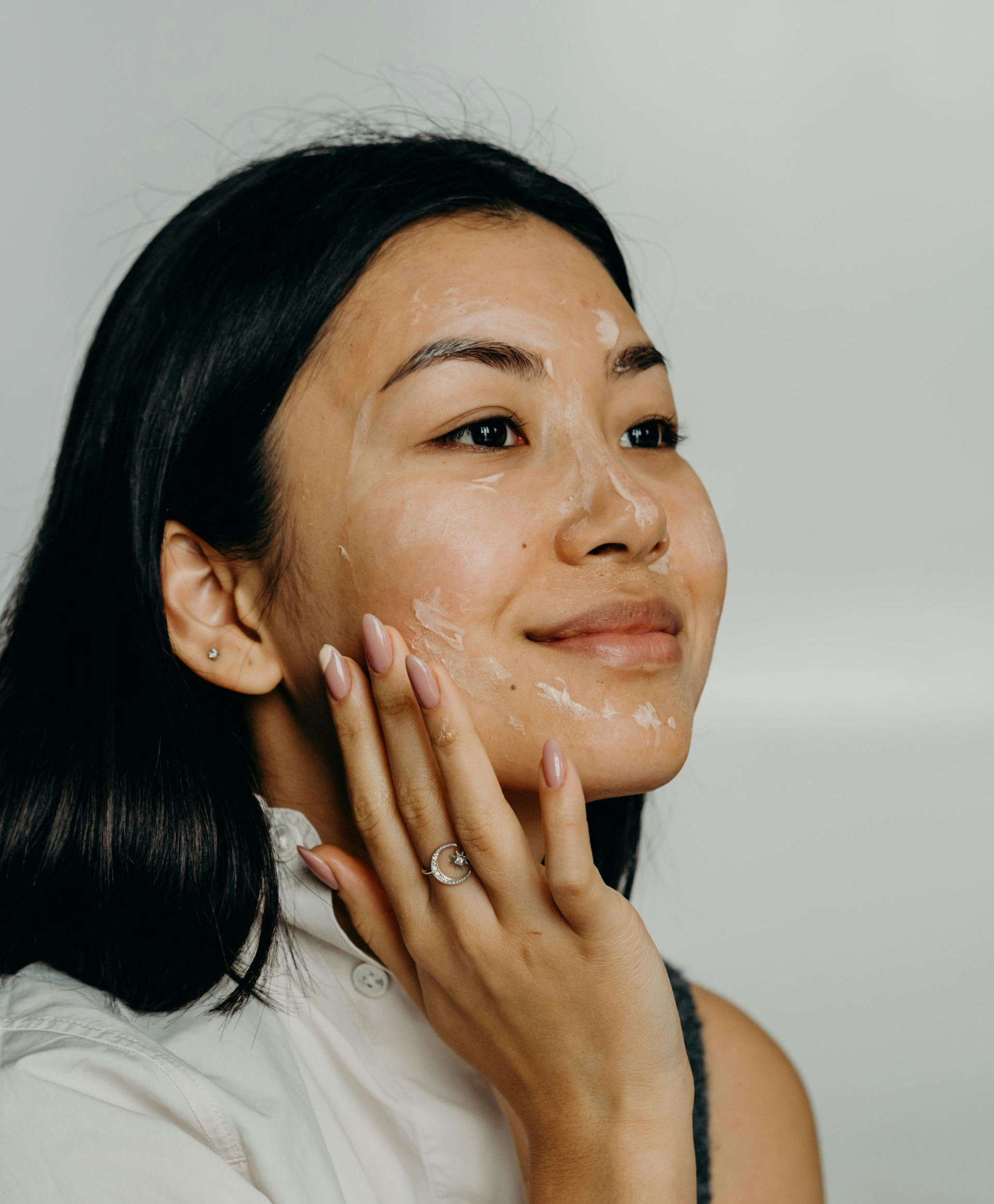 Een persoon met donker haar en een neutrale uitdrukking brengt voorzichtig met de vingertoppen crème aan op zijn of haar wang.