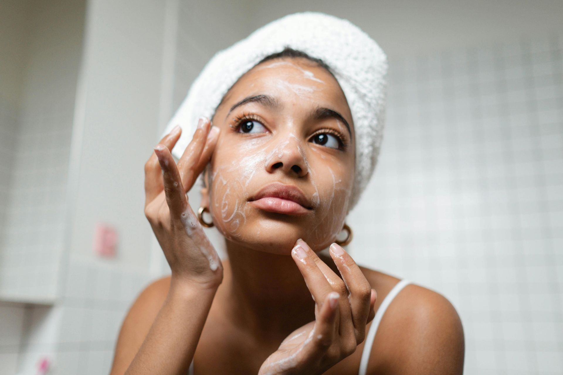 A person with a white towel wrapped around their head gently applies skincare product to their face in a bathroom.