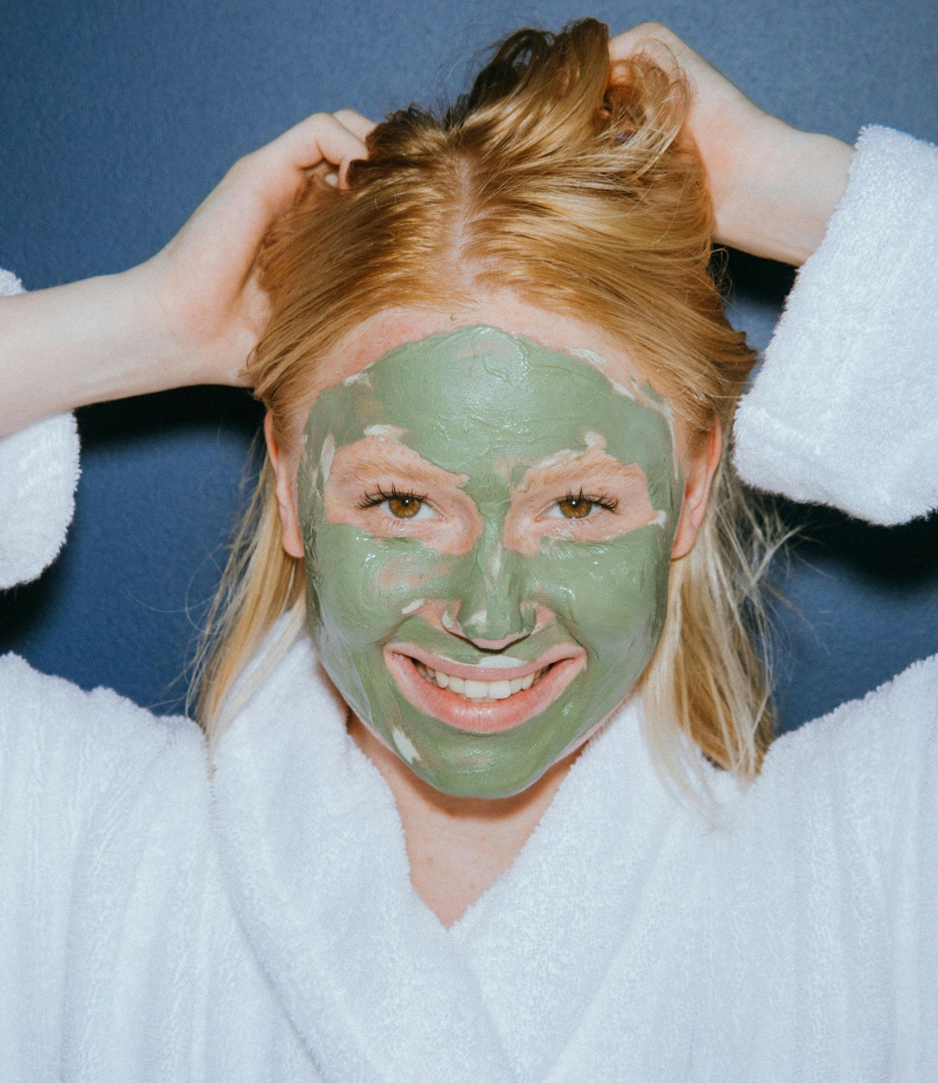 Woman with green face mask, holding hair up, smiling, wearing a white robe.