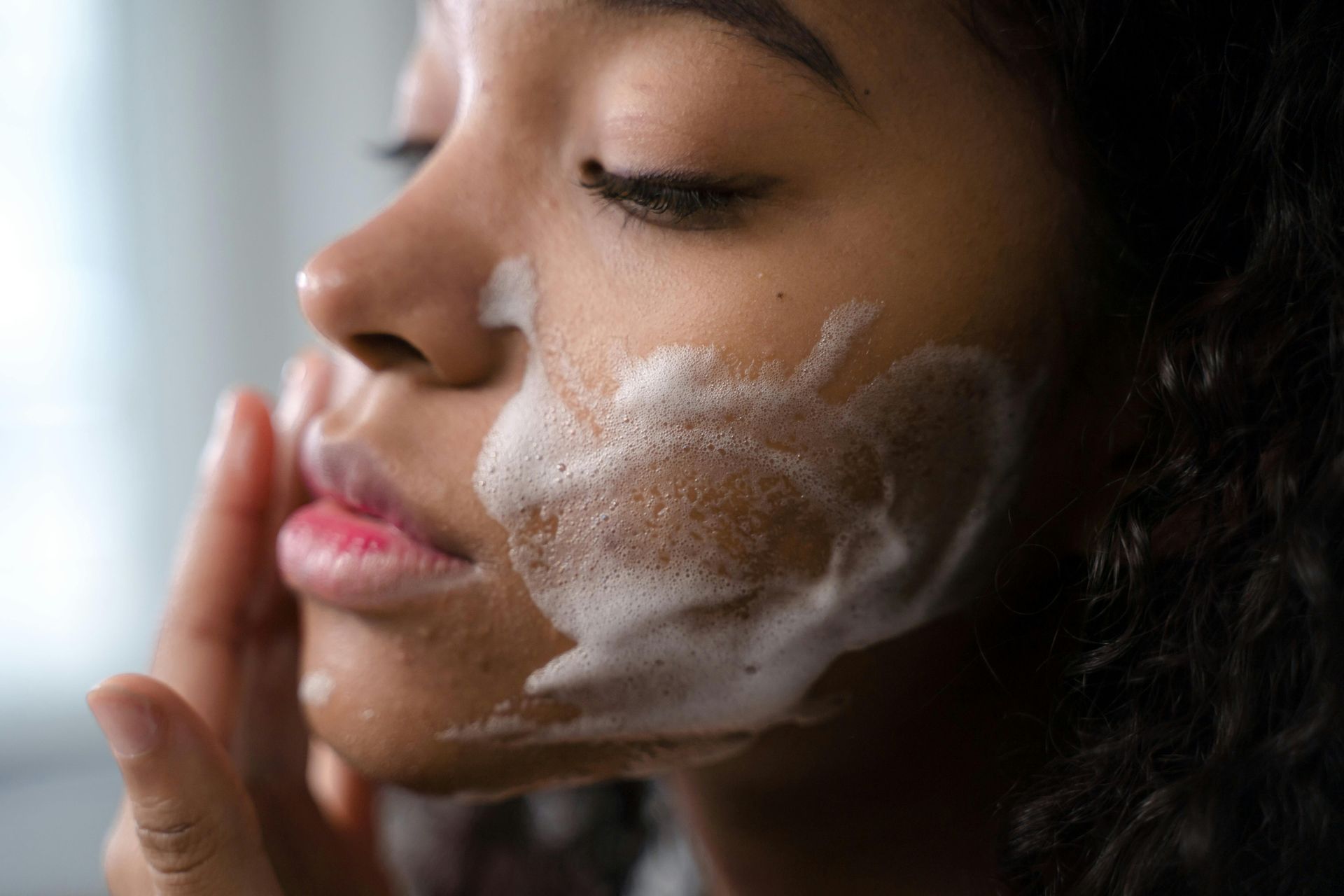Woman with closed eyes, applying foamy cleanser to her cheek.