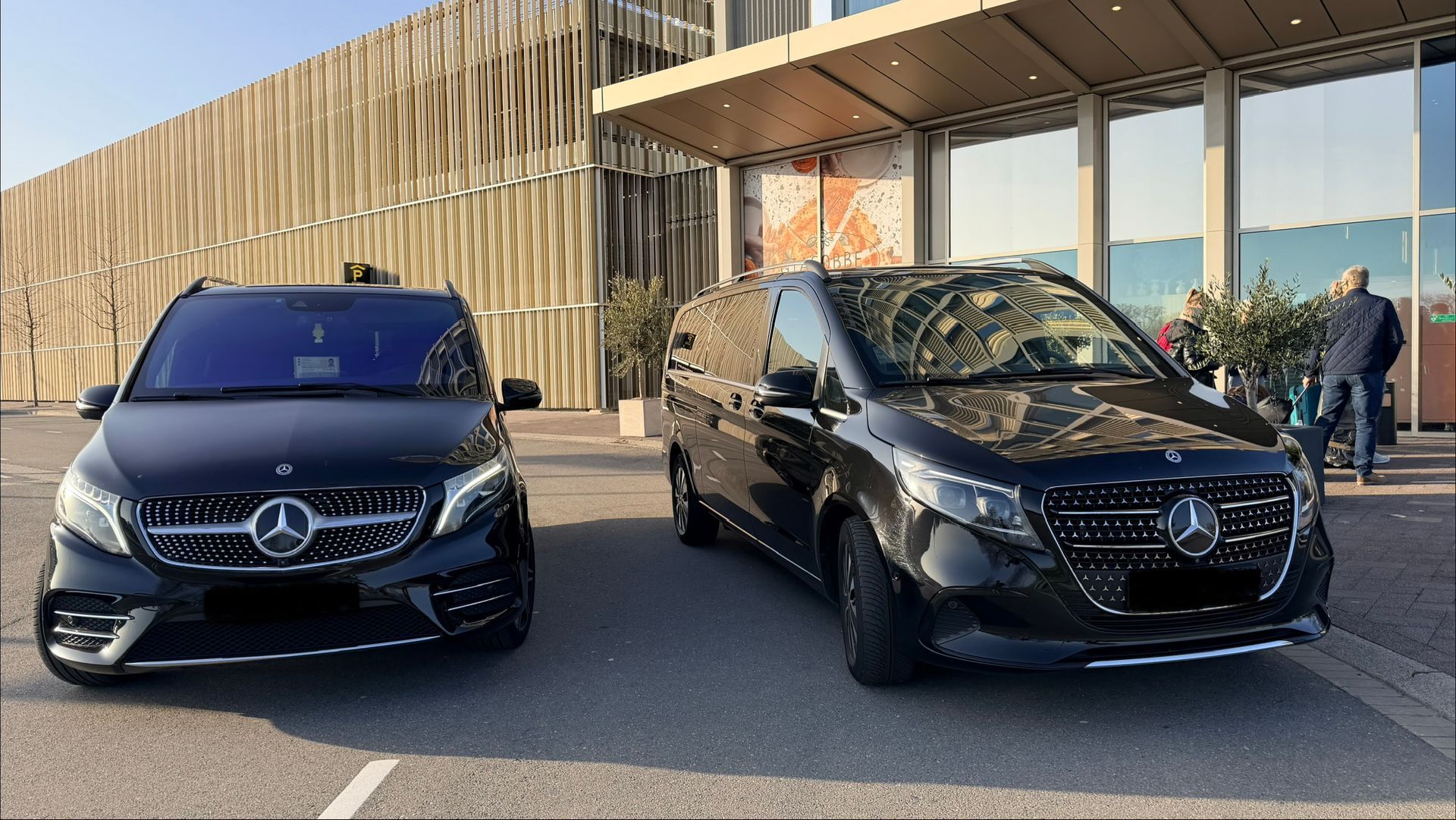 Two black Mercedes-Benz vans parked side-by-side in front of a modern building with a canopy 