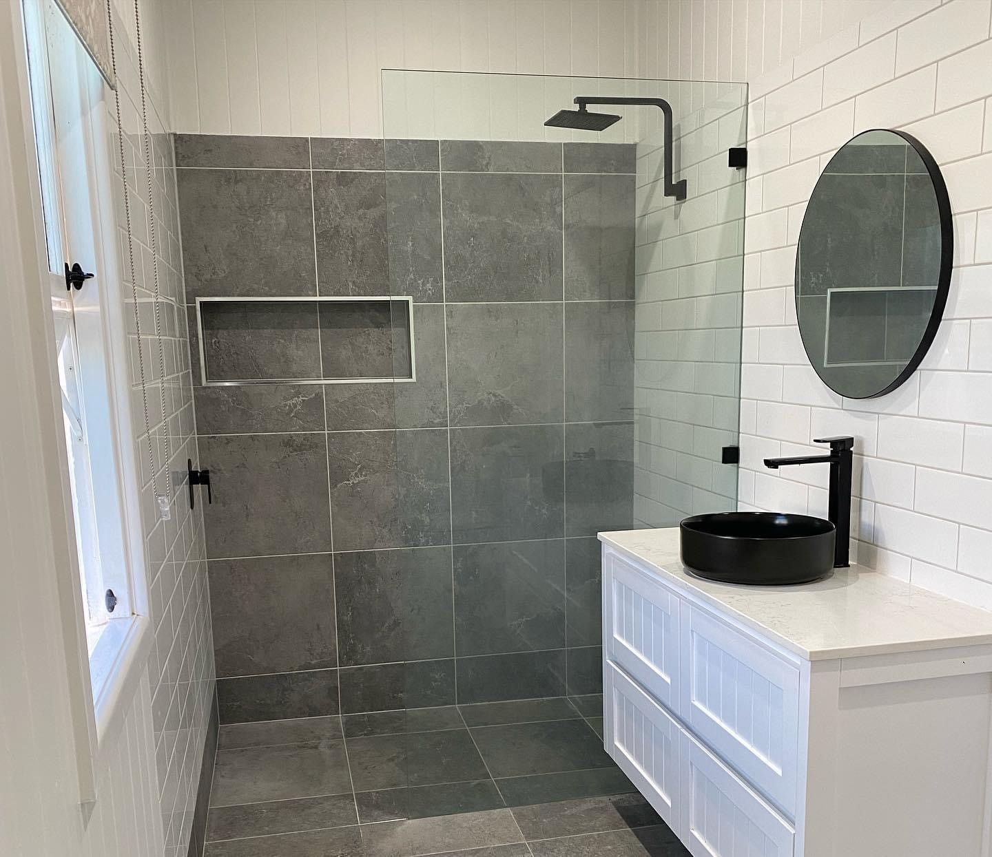 Modern bathroom shower with gray tile, white vanity, round mirror, and black sink faucet. 