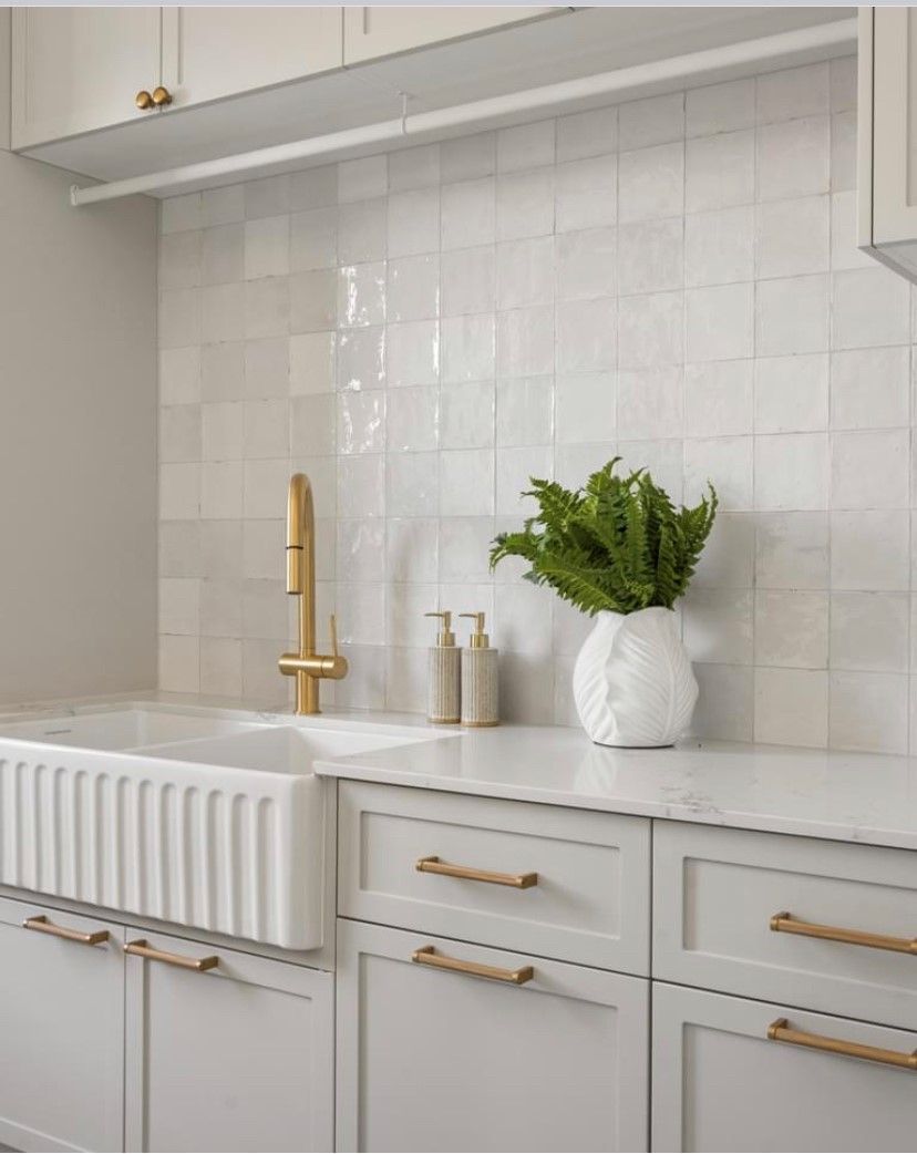 White farmhouse-style sink and cabinets with gold fixtures and green foliage in a well-lit space.