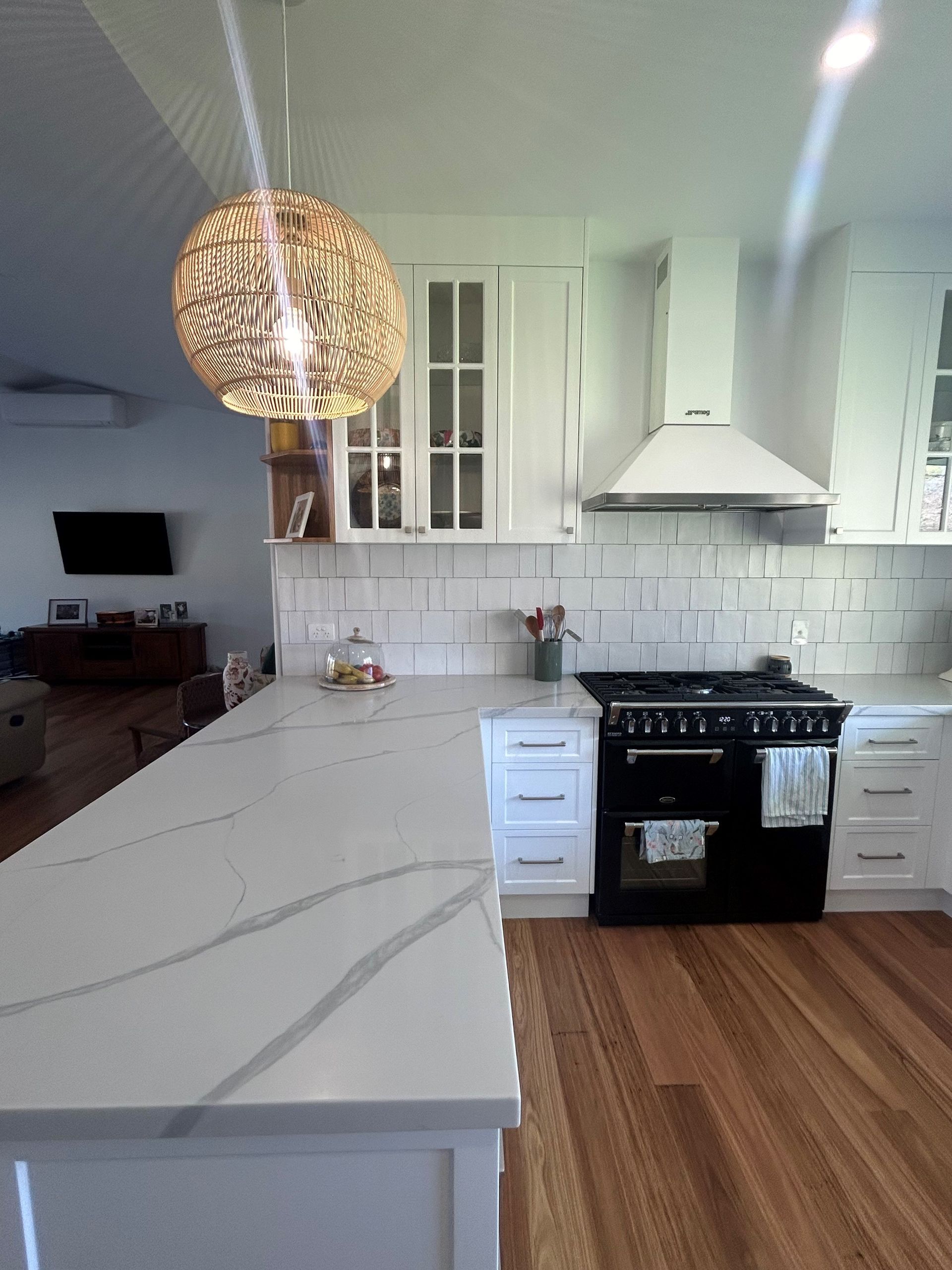 Bright white kitchen with marble island, black stove, wood floor, and woven pendant light — Valley Cabinetmakers In Biloela, QLD 