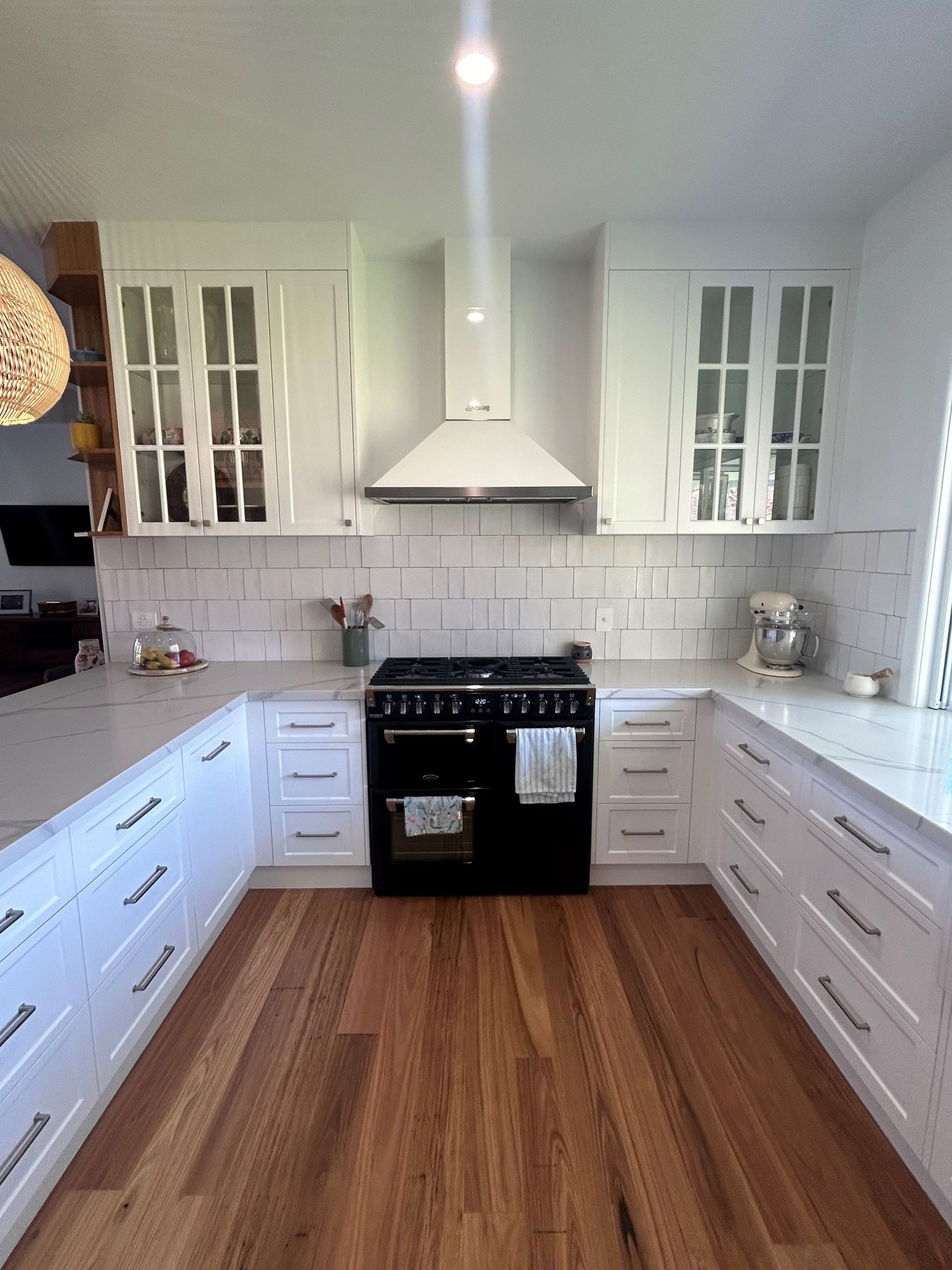 Bright white kitchen with black stove, long cabinets, glass-front uppers, and wood floor. — Valley Cabinetmakers In Biloela, QLD 