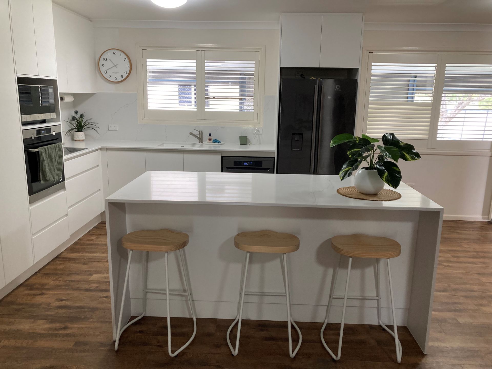 Bright white kitchen with island, three stools, black fridge, wall clock, and potted plant — Valley Cabinetmakers In Biloela, QLD 