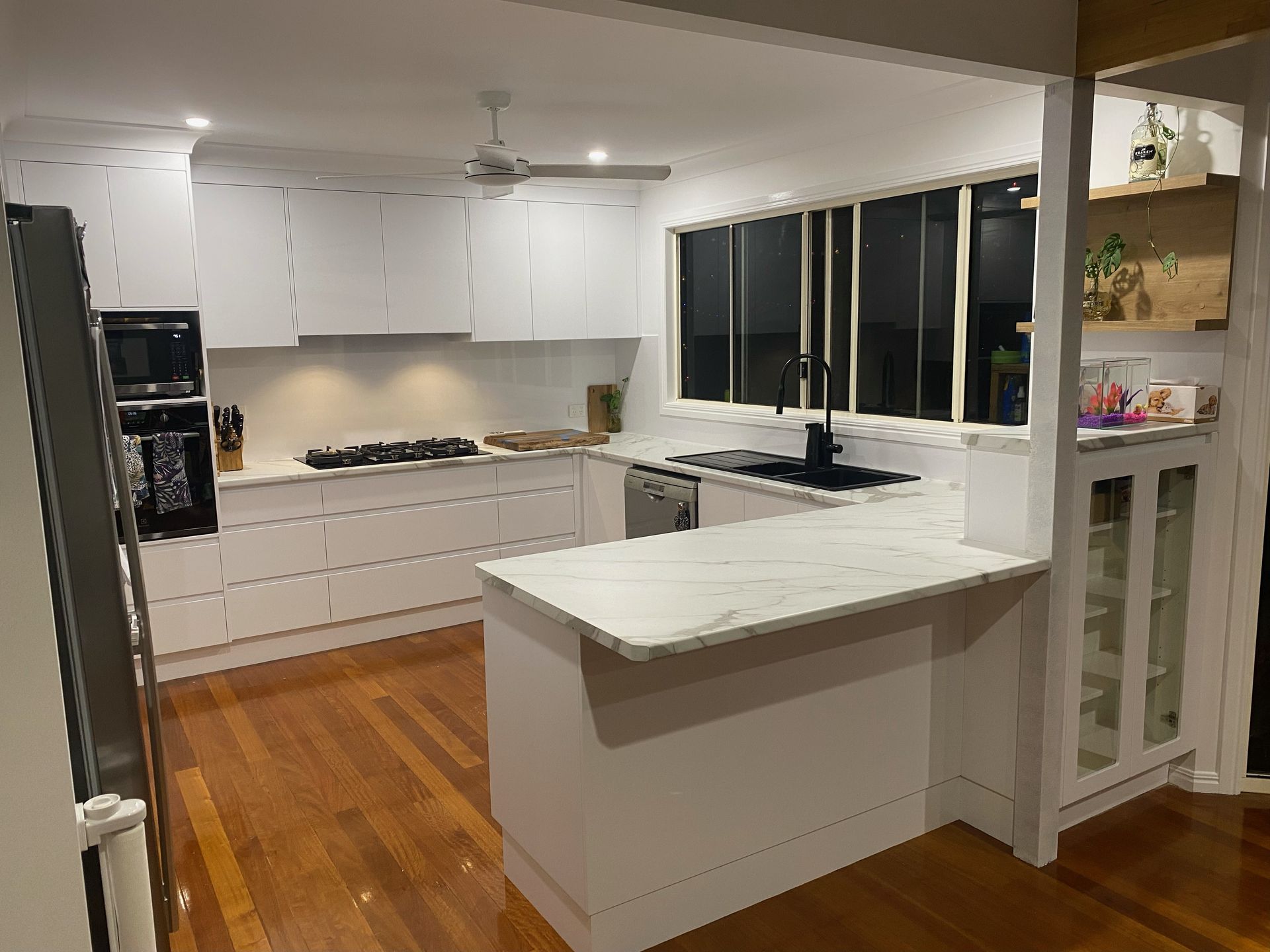 Modern white kitchen with island, wood floors, black countertops, and large windows at night — Valley Cabinetmakers In Biloela, QLD 