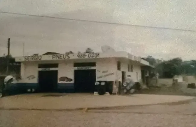 Pequena loja à beira da estrada com fachada branca e azul, motocicletas estacionadas e uma rua de terra sob um céu nublado.