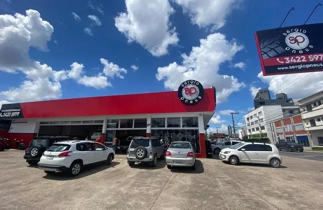 Estacionamento em frente a uma concessionária com telhado vermelho, com vários carros estacionados sob um céu azul nublado.