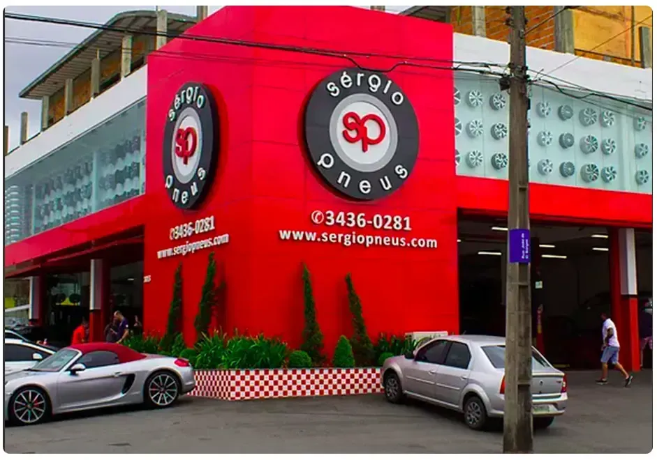 Fachada vermelha com logotipo circular, carros estacionados e pedestres em uma esquina.