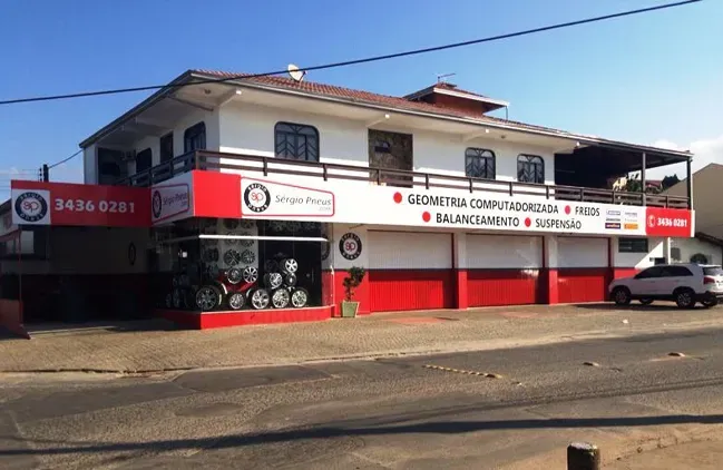 Prédio comercial de dois andares com detalhes em vermelho e placas na fachada, visto do outro lado da rua.