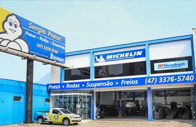 Fachada azul de uma oficina mecânica Michelin com placas de serviço e um Volkswagen amarelo estacionado em frente.