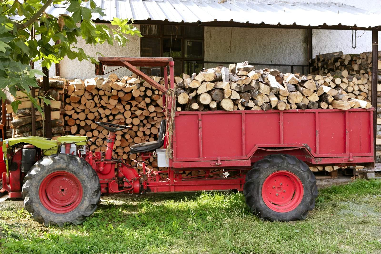 A tractor with a trailer full of logs is parked in front of a pile of logs.