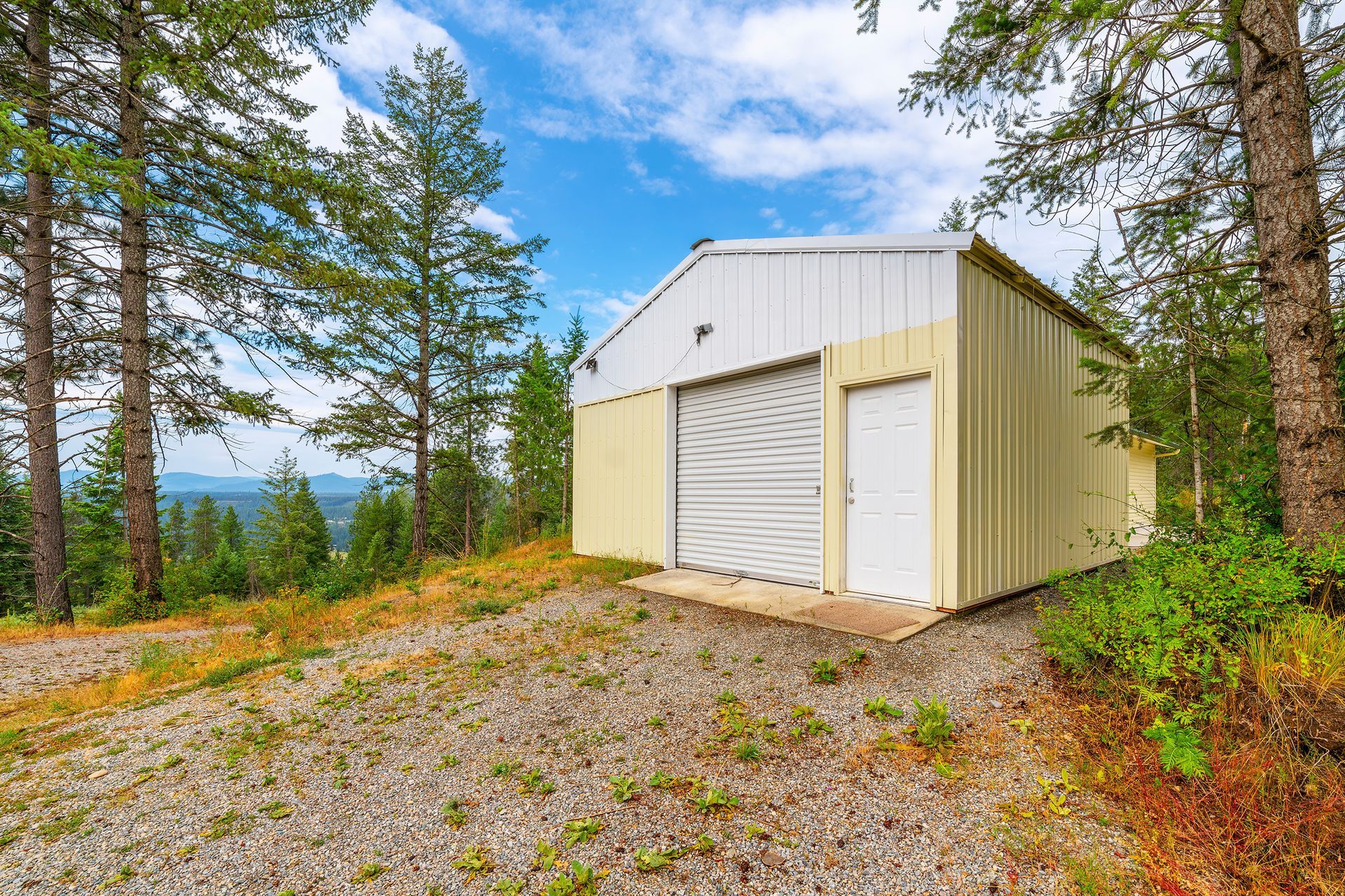 A large metal garage is sitting on top of a gravel hill surrounded by trees.