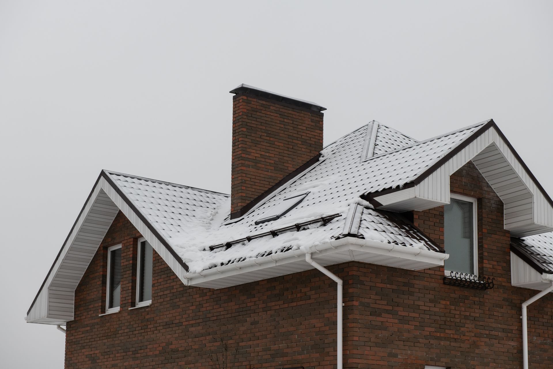 Brick house roof covered in snow; chimney; white trim; overcast sky.
