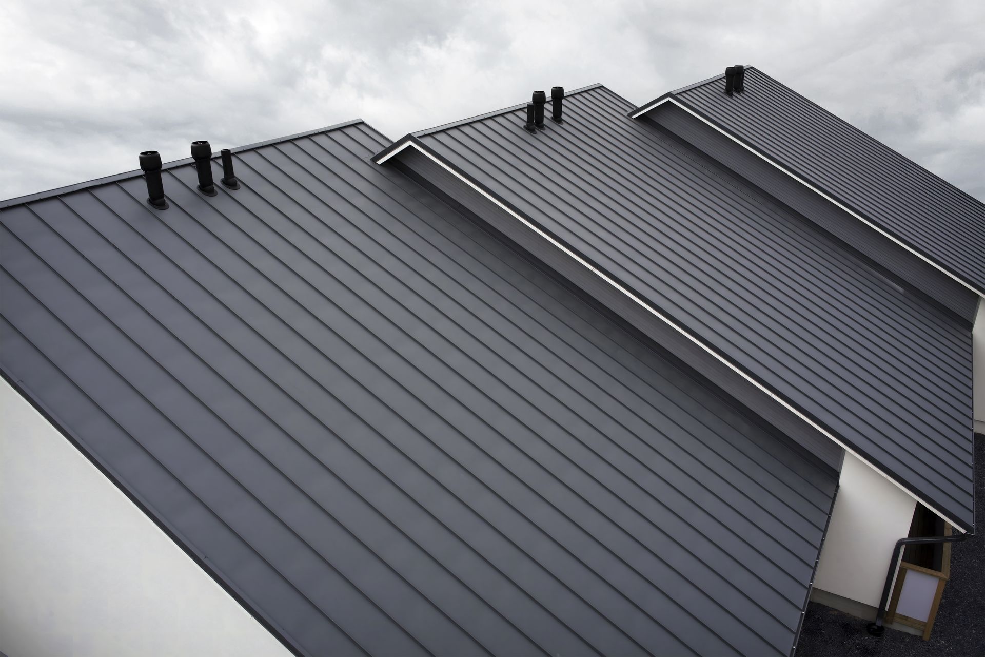 Dark gray metal roof on a building with three sections, cloudy sky.