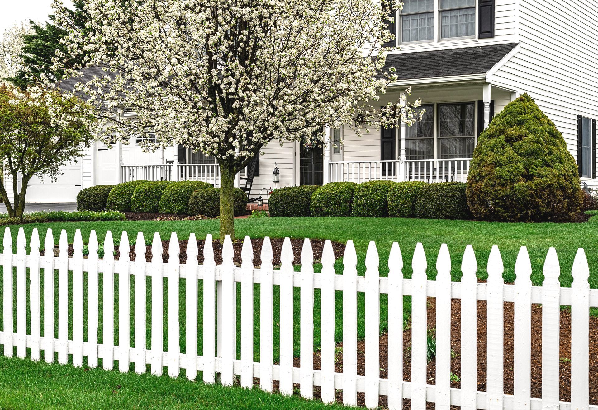 A white picket fence surrounds a lush green lawn in front of a house.