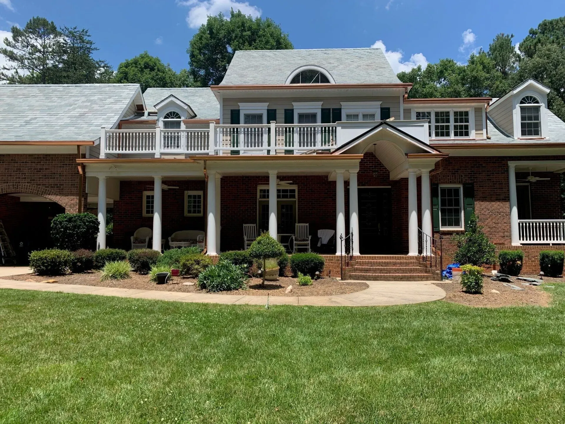 A large brick house with a large porch and stairs is sitting on top of a lush green lawn.