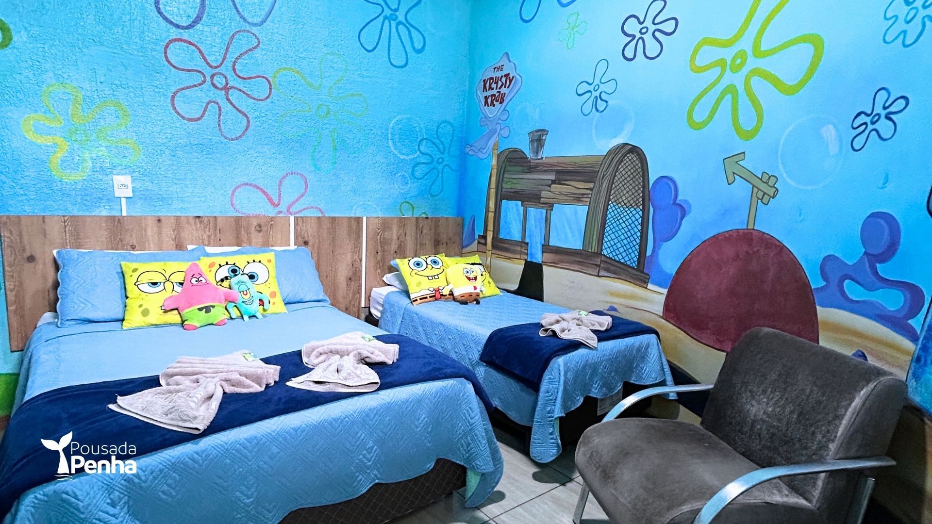 Um quarto de hotel temático do Bob Esponja com duas camas azuis, murais florais nas paredes e uma poltrona cinza.