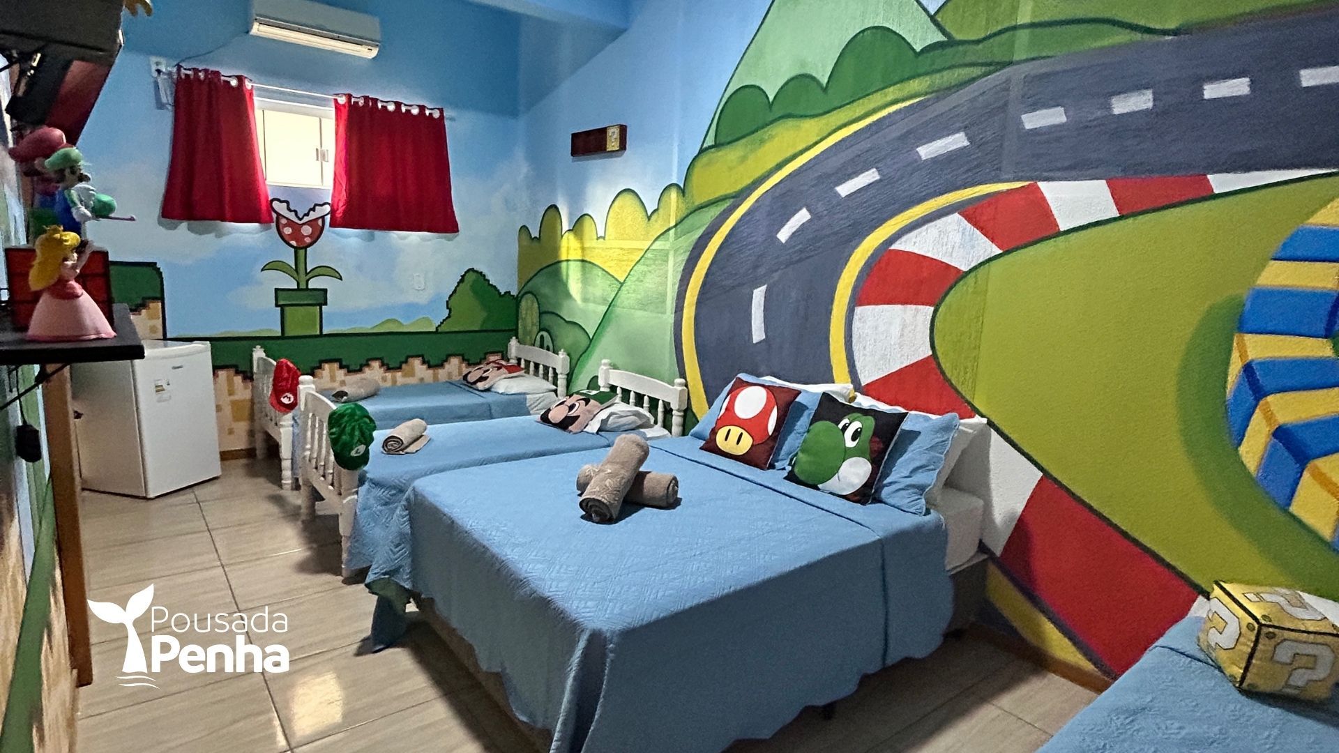 Um quarto temático do Super Mario com um mural de pista de corrida na parede, três camas azuis e almofadas temáticas.