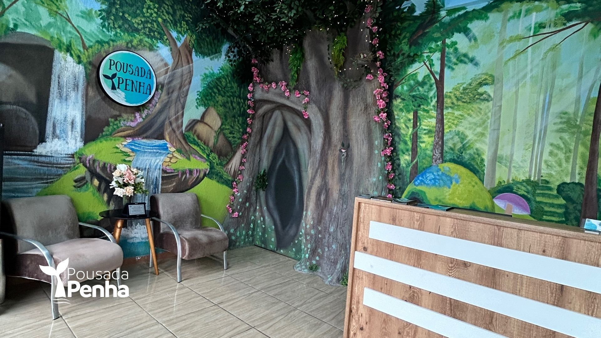 Área de espera interna com cadeiras em frente a um mural pintado representando uma floresta, uma grande estrutura em forma de árvore e um balcão de recepção de madeira.