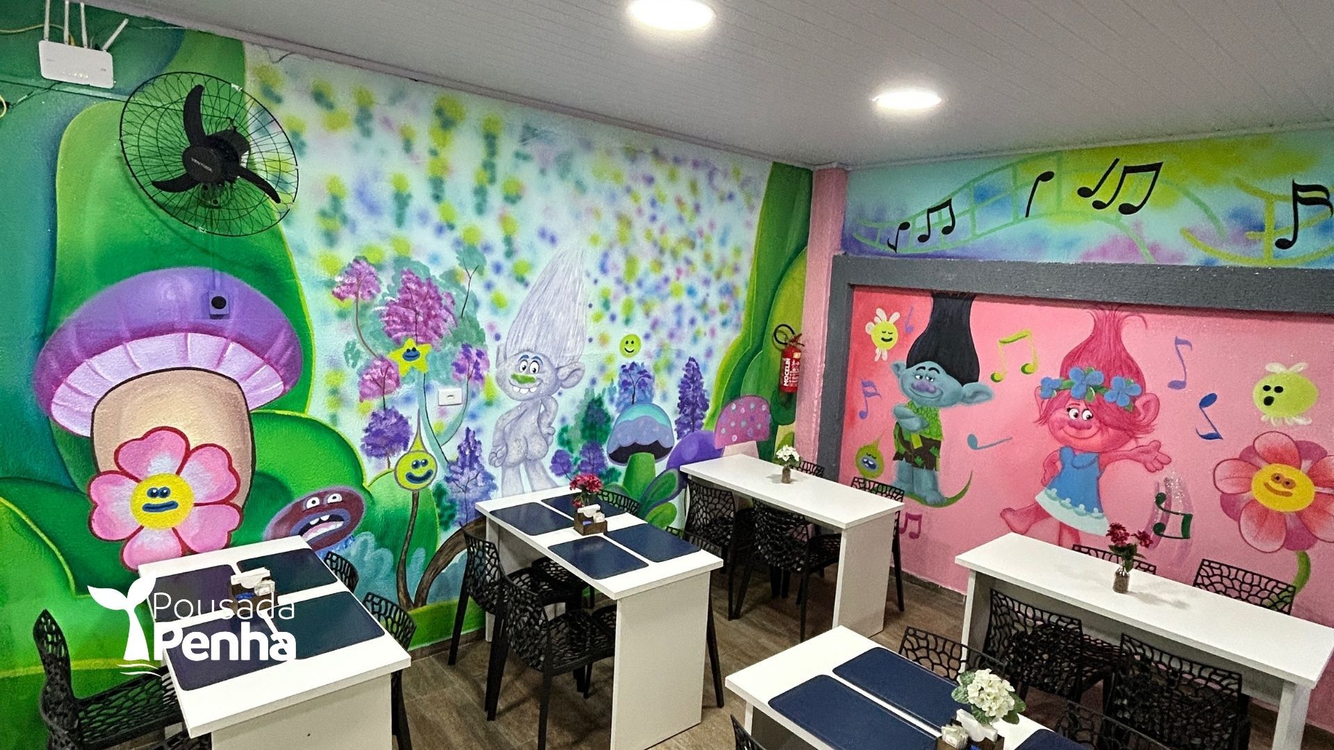 Pequena sala de jantar decorada com personagens dos Trolls, murais coloridos nas paredes, mesas brancas e cadeiras pretas.