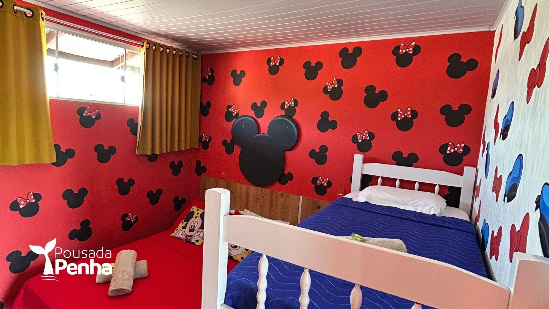 Um quarto decorado com motivos do Mickey e da Minnie Mouse, com paredes vermelhas com silhuetas pretas, duas camas e cortinas.