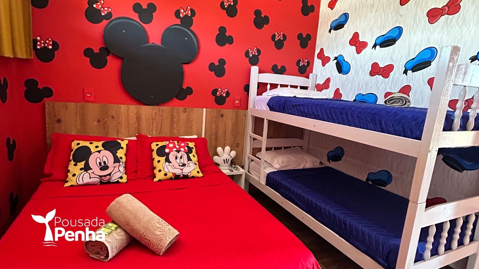 Um quarto temático com o Mickey e a Minnie Mouse, apresentando uma cama vermelha, um beliche branco com roupa de cama azul e paredes estampadas.