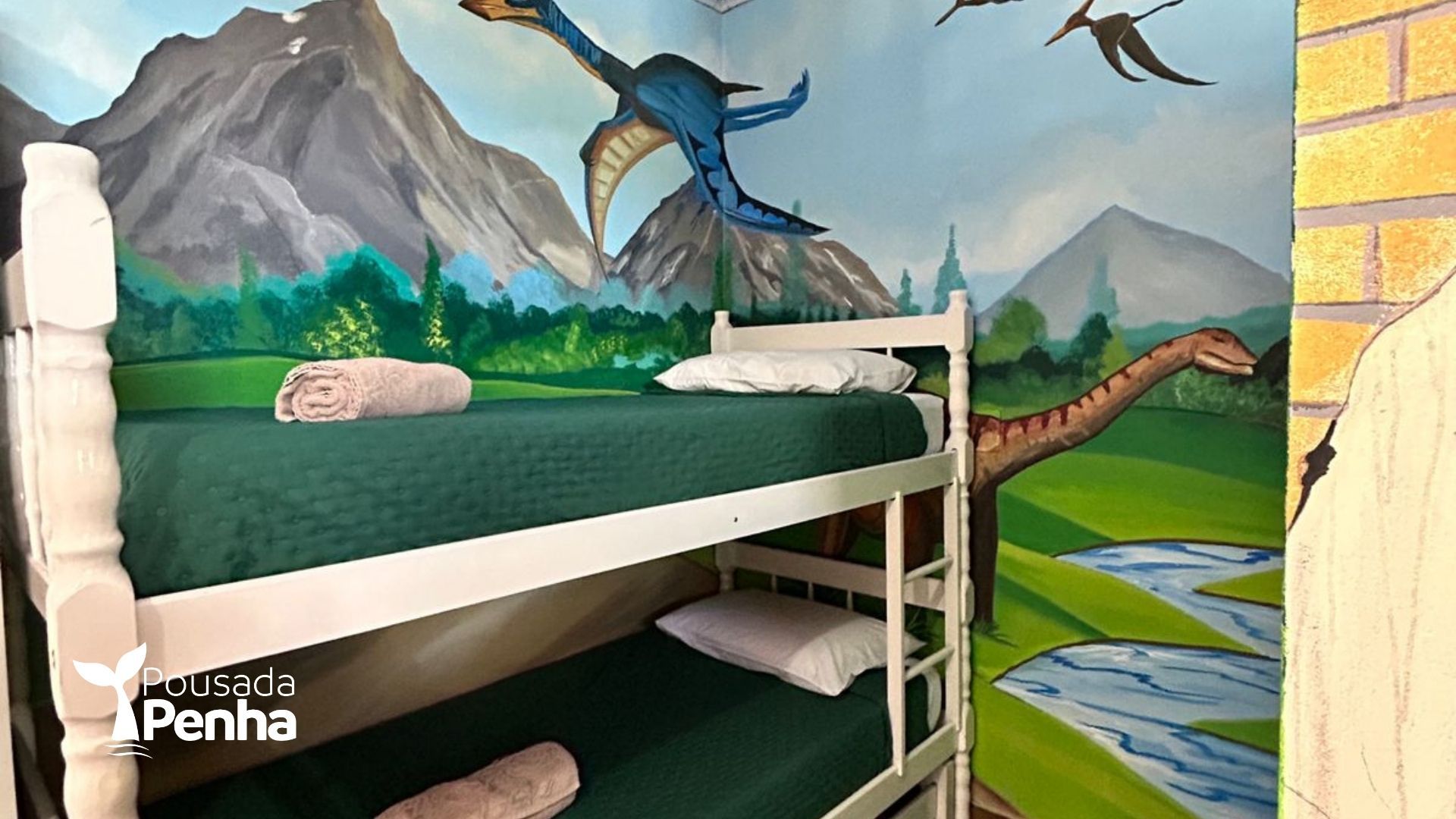 Uma beliche com estrutura branca e roupa de cama verde em um quarto com um mural na parede representando montanhas, um rio e dinossauros.
