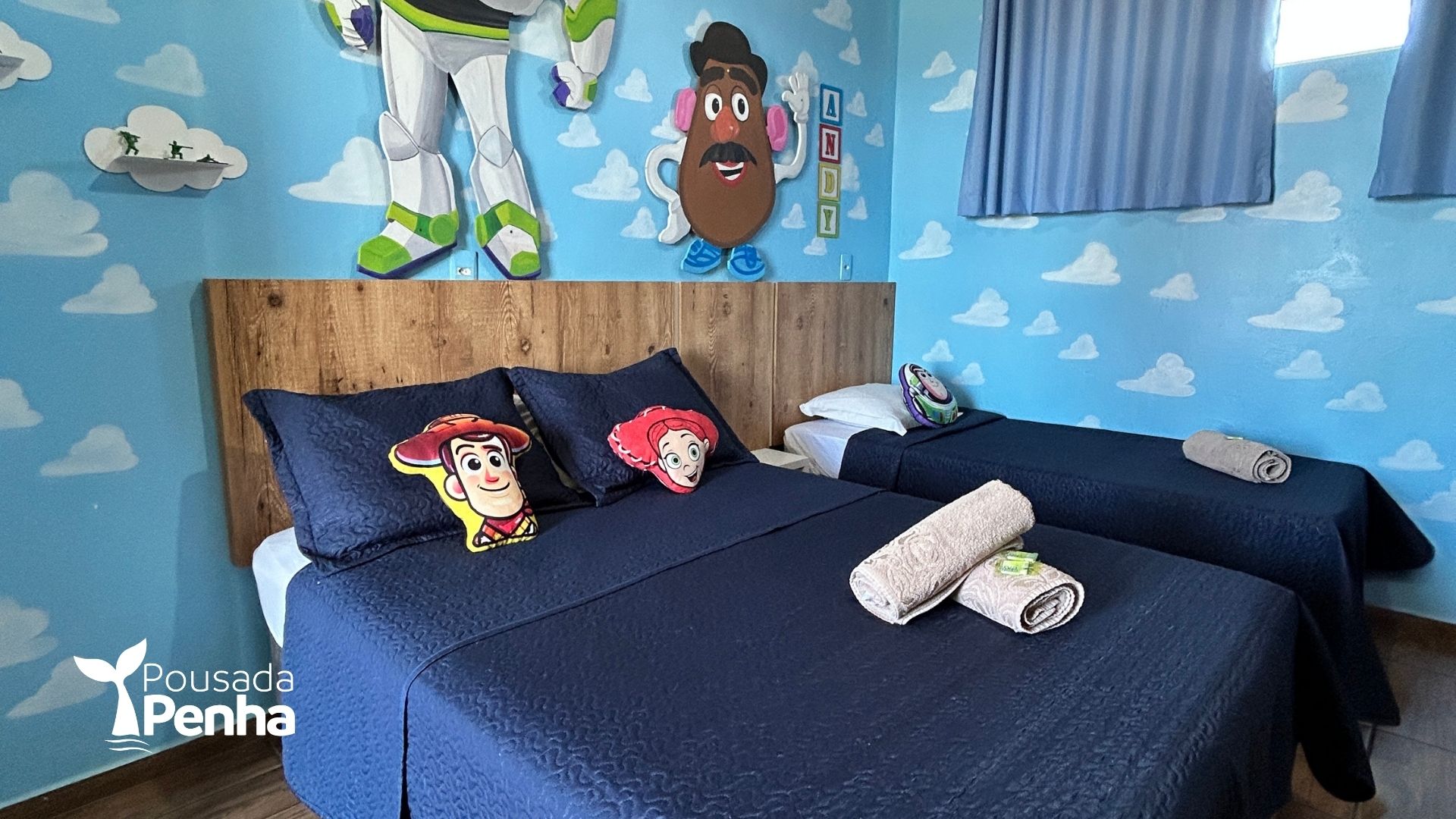 Quarto temático de Toy Story com paredes estampadas com nuvens, duas camas com roupa de cama azul-escura e adesivos de parede com os personagens.
