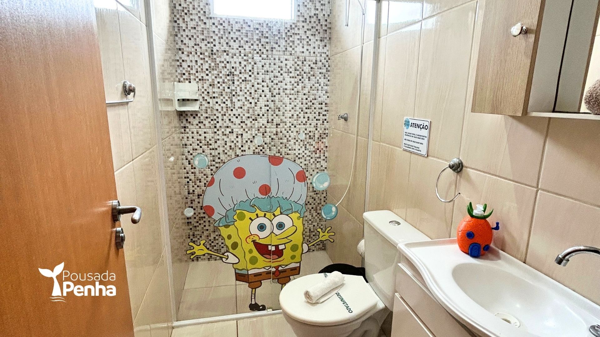 Banheiro com cortina de chuveiro do Bob Esponja, paredes revestidas com azulejos bege, vaso sanitário, pia e decoração em formato de abacaxi.