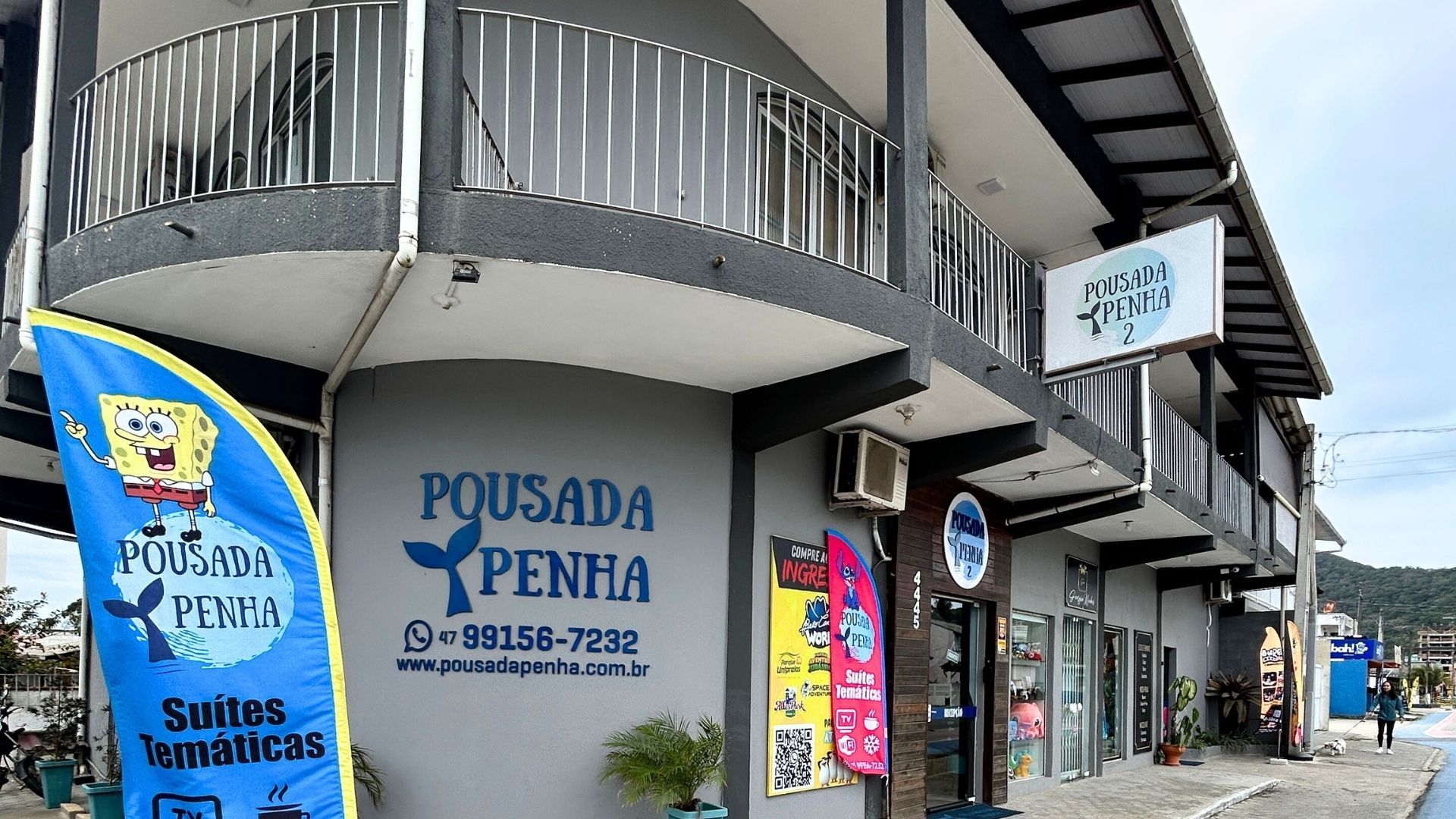 Vista externa da Pousada Penha, um prédio cinza de dois andares com uma bandeira azul do Bob Esponja, sinalização e uma varanda.