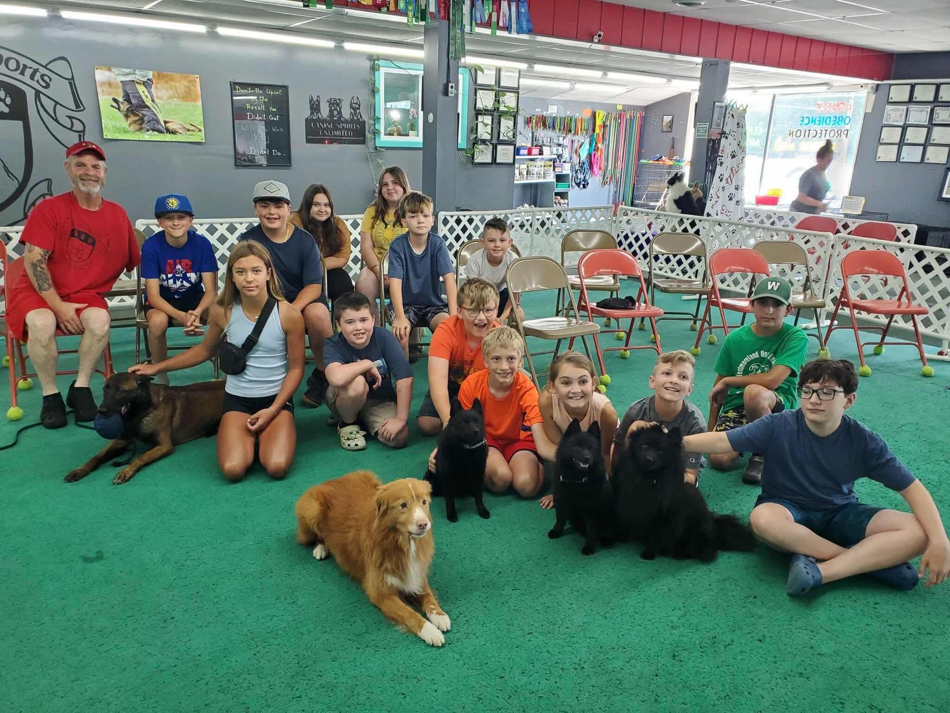 A group of children and two adults sitting with dogs indoors. They appear to be in a dog training facility.