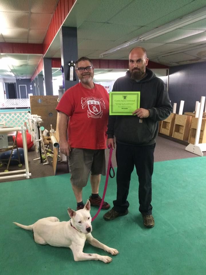 Two men and a white dog pose with a certificate inside a building. The dog is lying on the floor. One man holds the certificate.