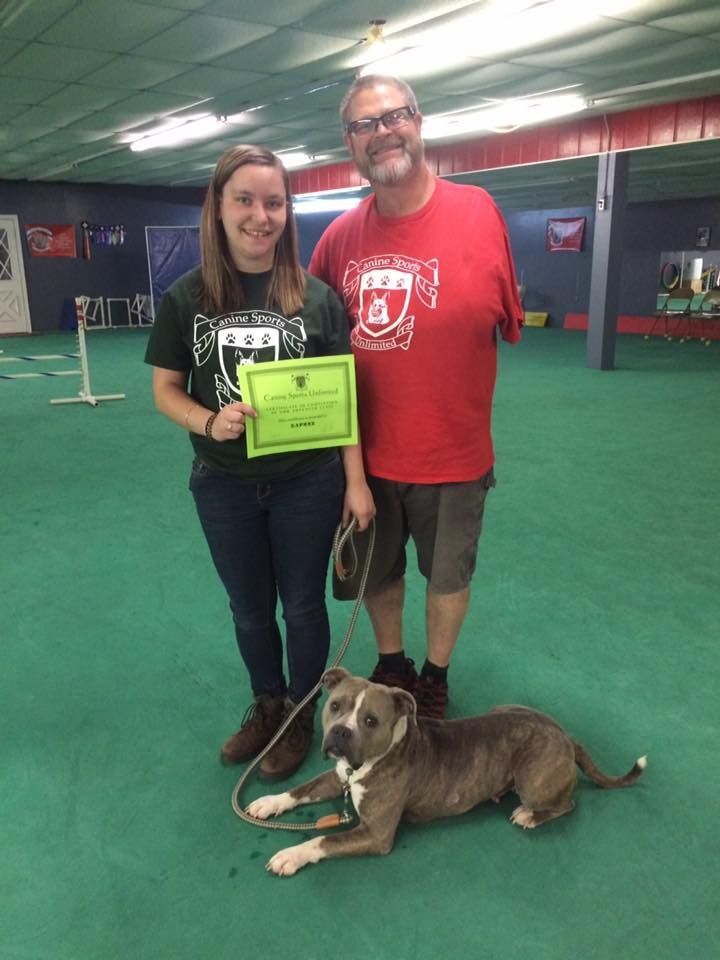 A young woman and a man stand with a dog, holding a certificate, on a green floor inside a training facility.