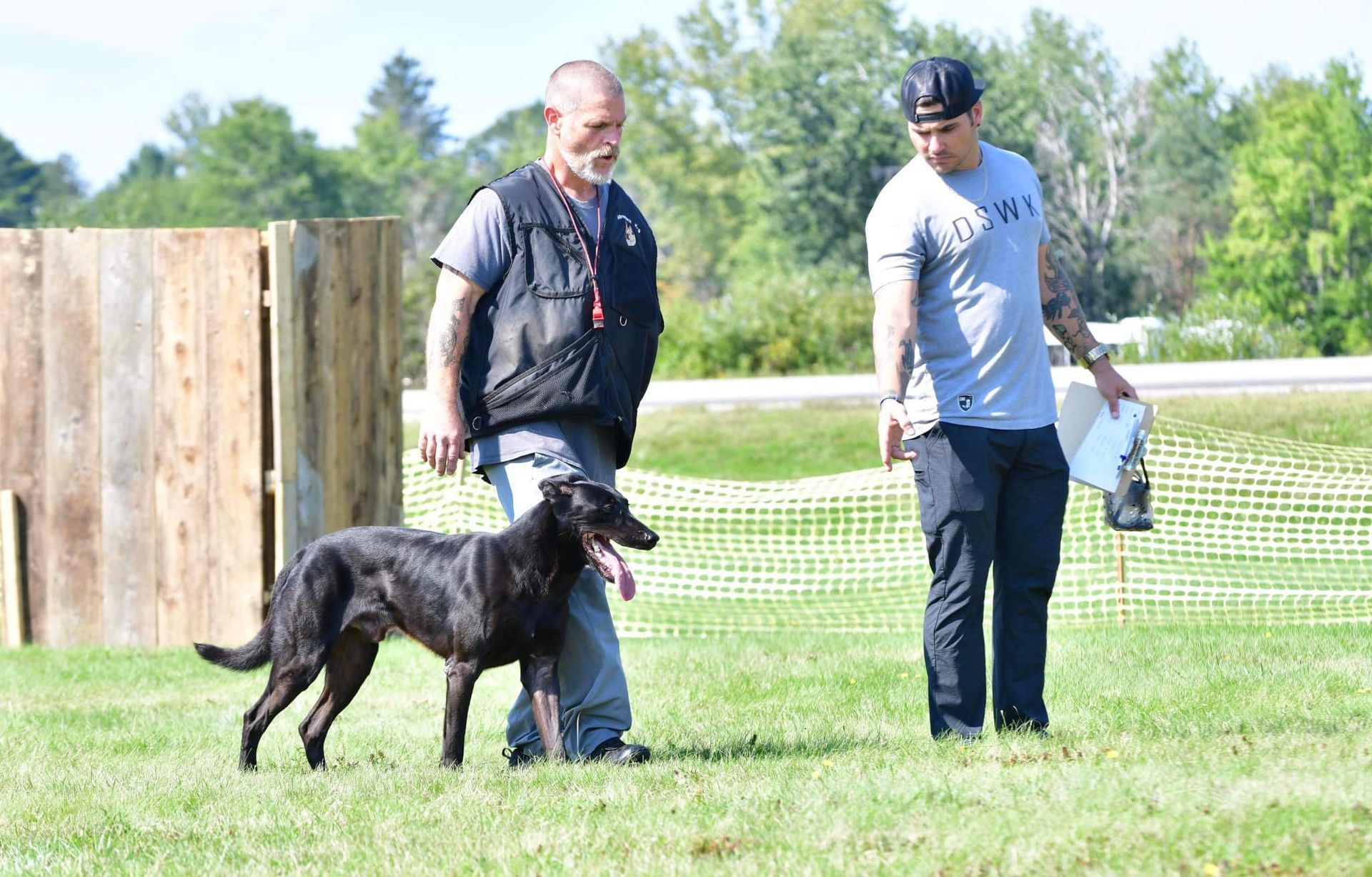 Two men train a black dog on a grassy field. One man holds a leash, while the other observes with papers.