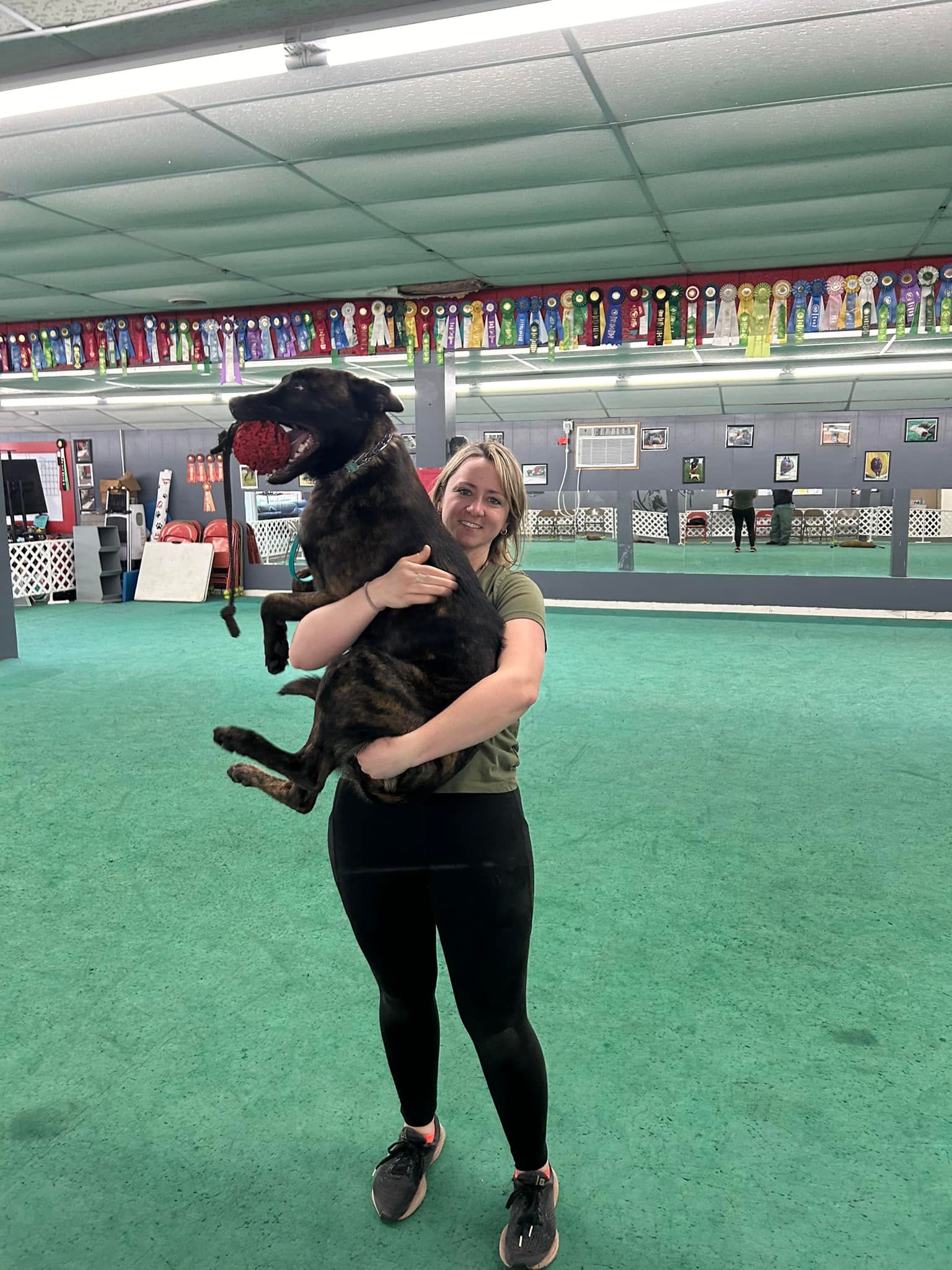 Woman holding a large, dark-colored dog in an indoor dog park. Both are smiling. The dog holds a red ball.