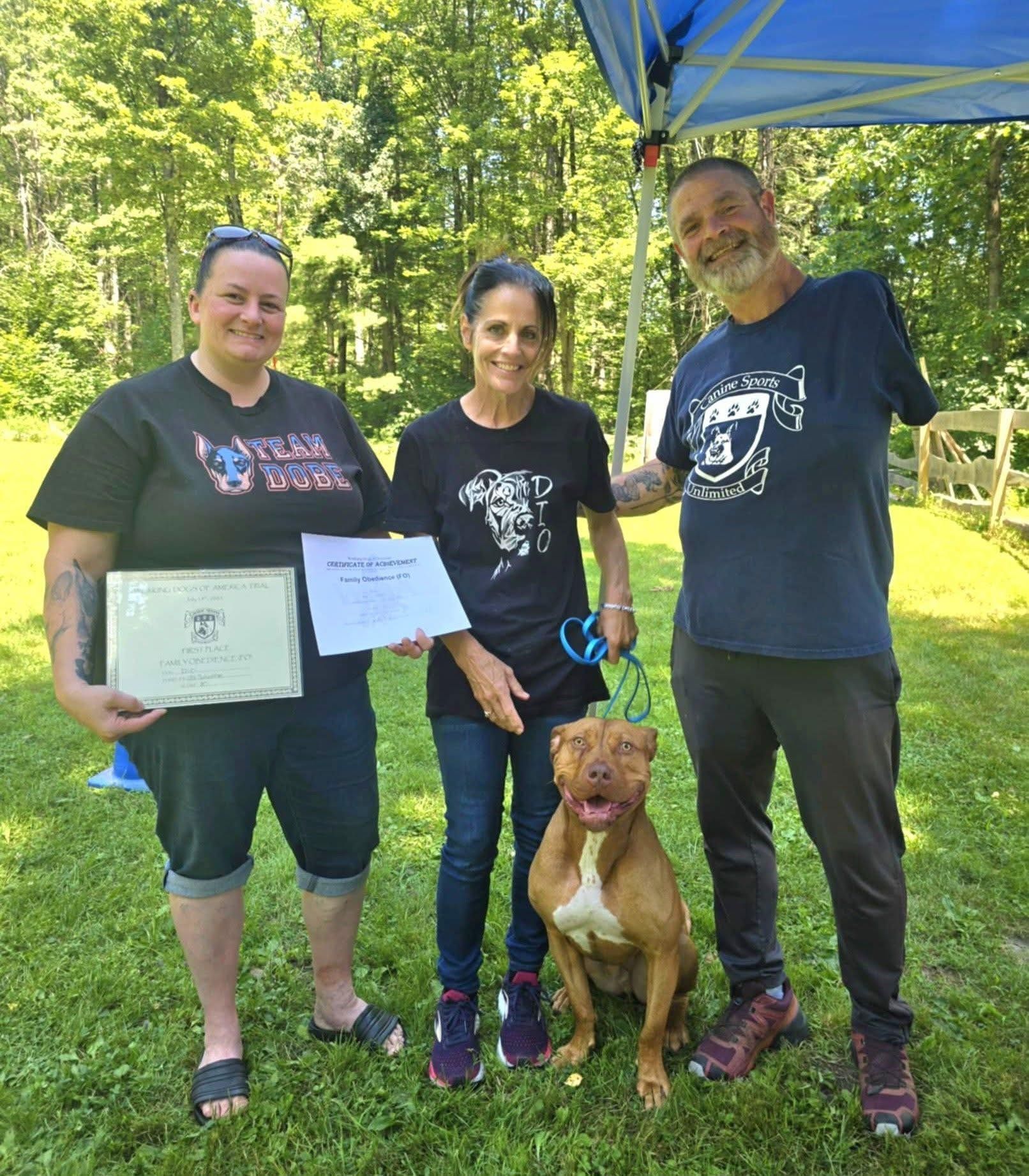 Three people and a brown dog pose outdoors on grass. Two women hold certificates; a man smiles, arm amputated.