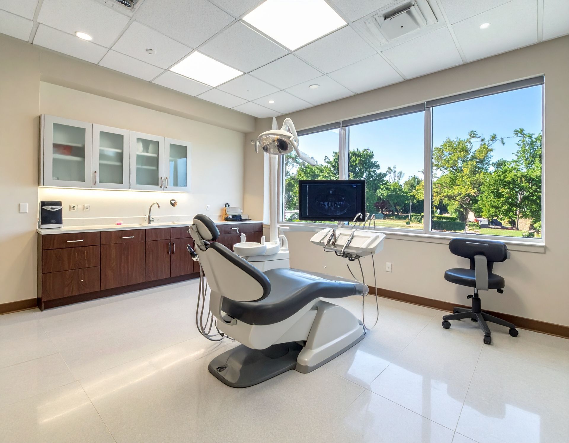Dental examination room with chair, equipment, and large window overlooking trees.