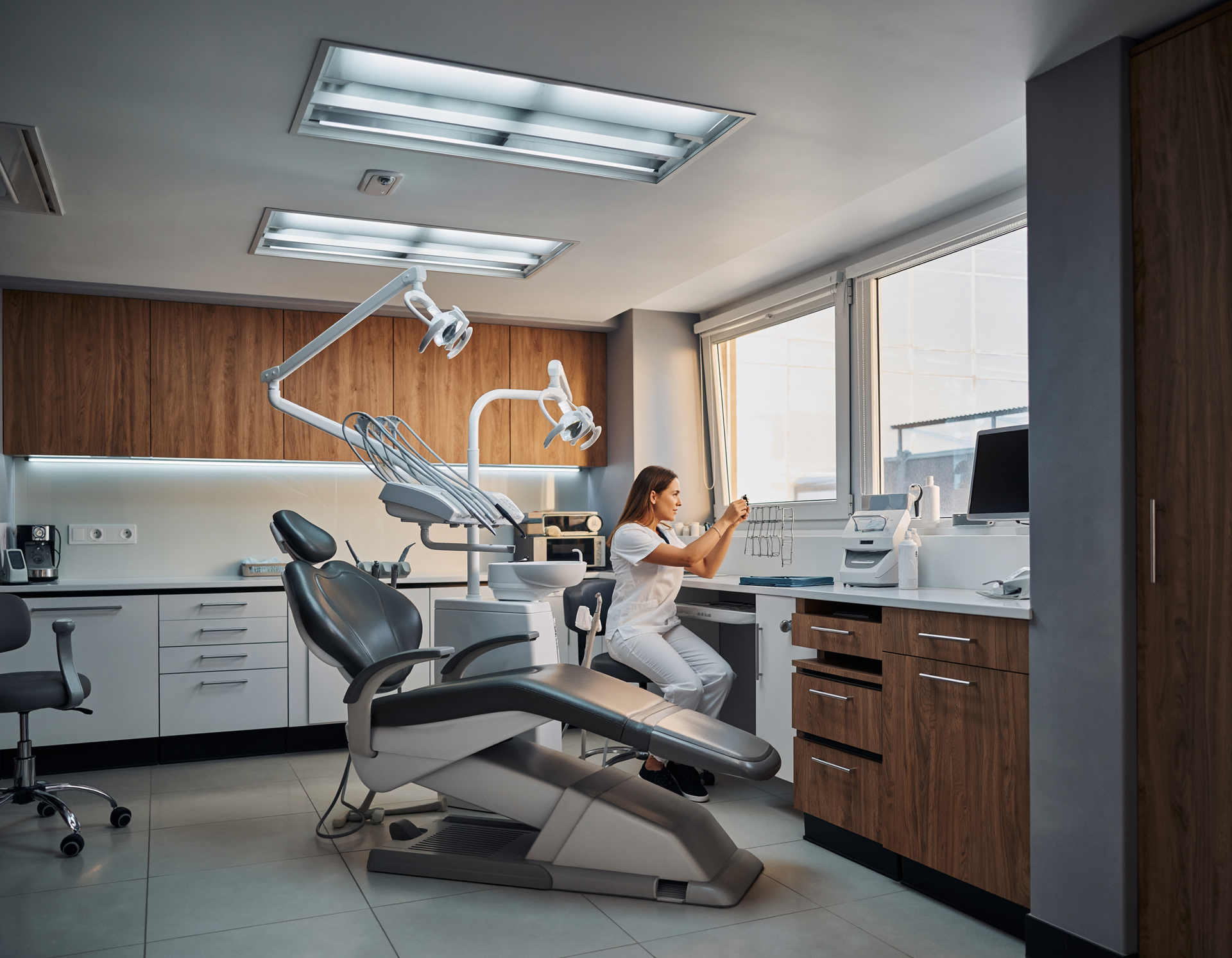 Dental office interior with a dentist adjusting equipment near a patient chair.