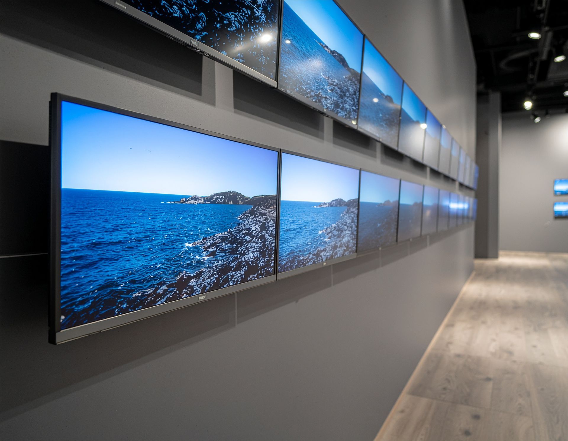 Gallery wall with multiple screens displaying a seascape. Blue water and rocky shoreline.
