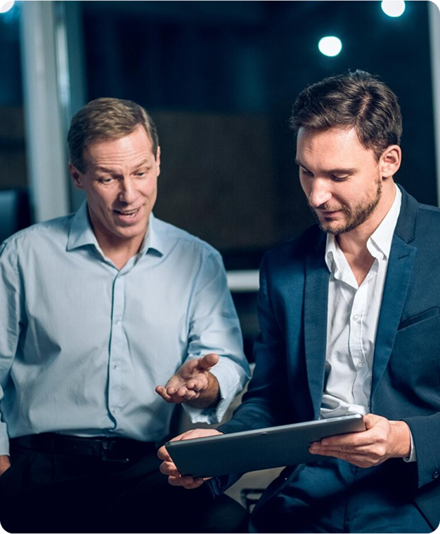Two men in business attire reviewing a tablet, one gesturing and talking.