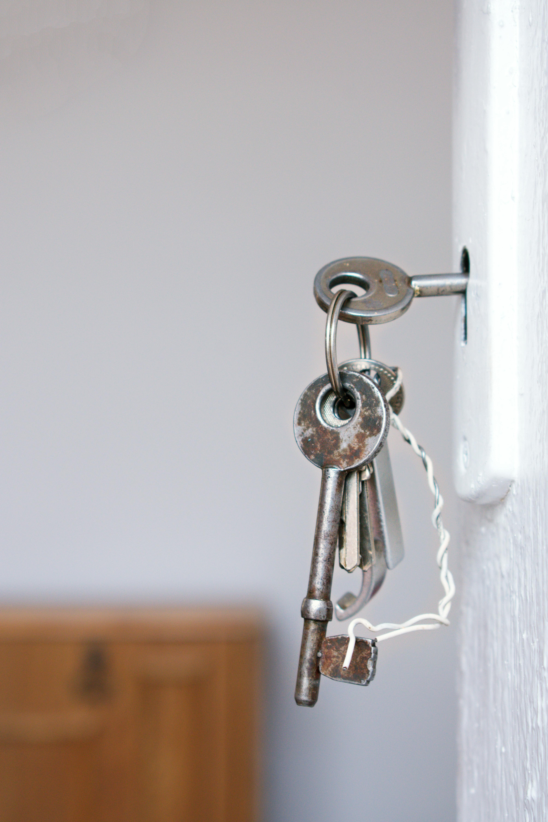Keys in a lock, hanging from a white door, with a wooden chest in the blurred background.