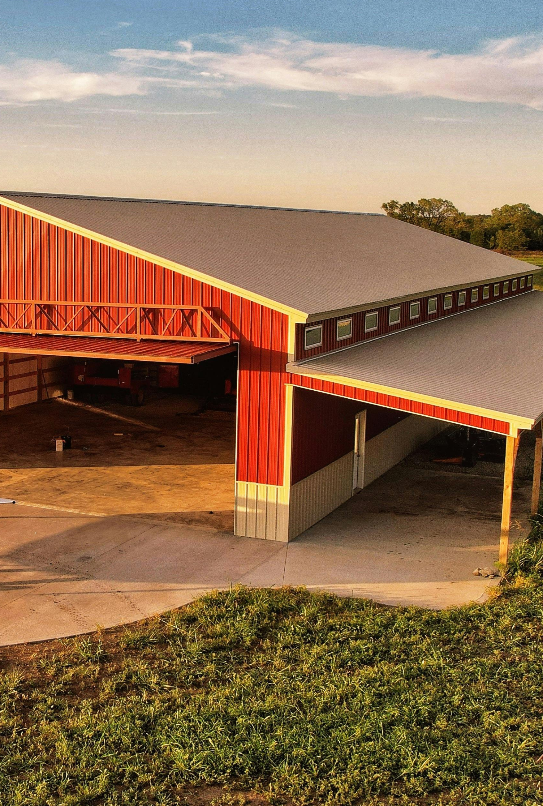 Red barn with a dark gray roof and a covered entryway; fields in the foreground and blue sky.