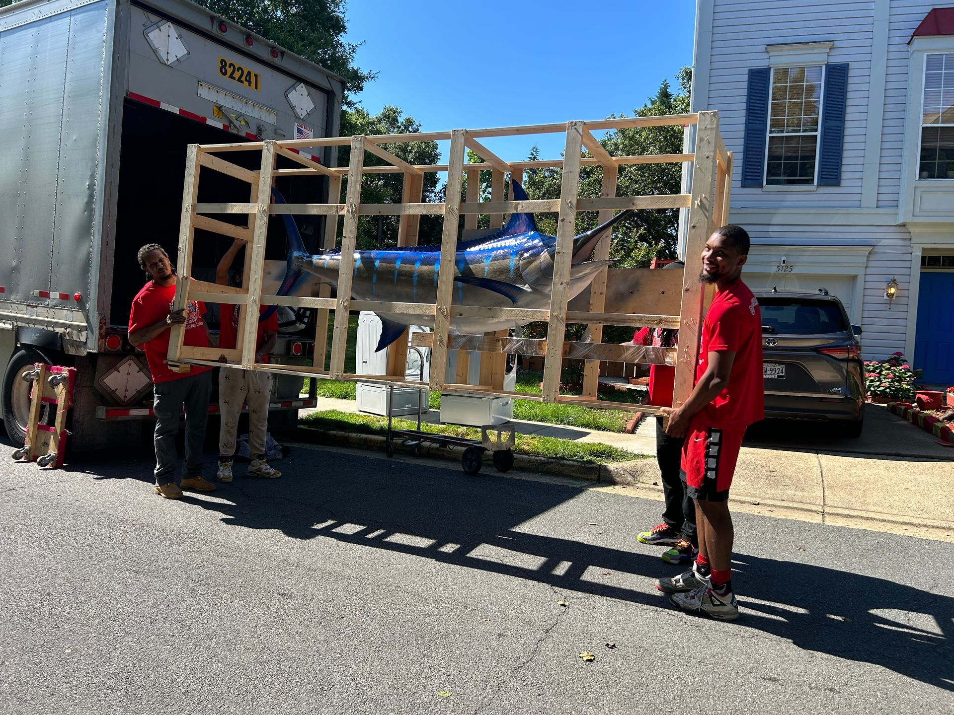 Movers loading a large, blue marlin sculpture in a wooden crate onto a moving truck.