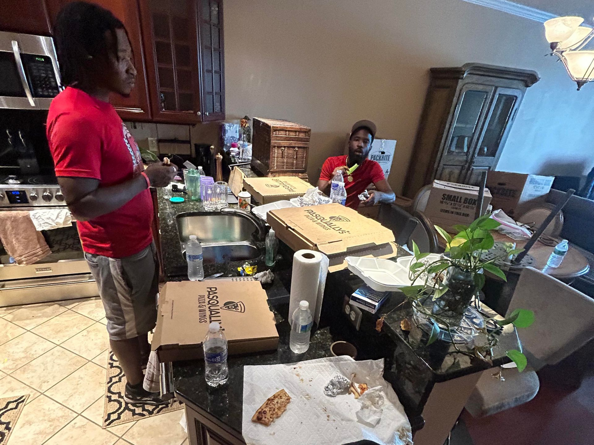 Two people in a messy kitchen with pizza boxes and trash. One person stands and the other sits.